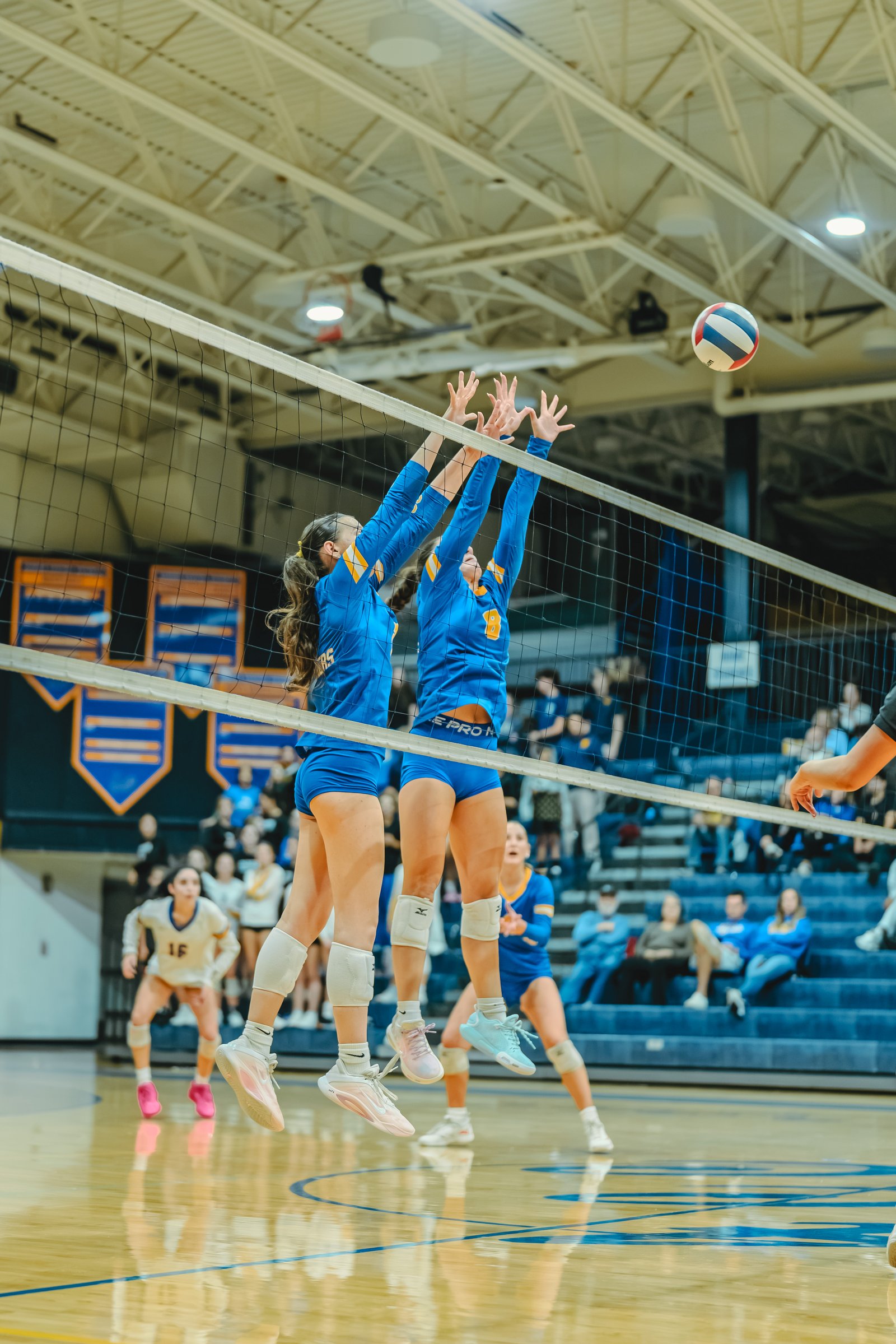 Players in blue jump to block a volleyball. A player in white with number 15 is visible.
