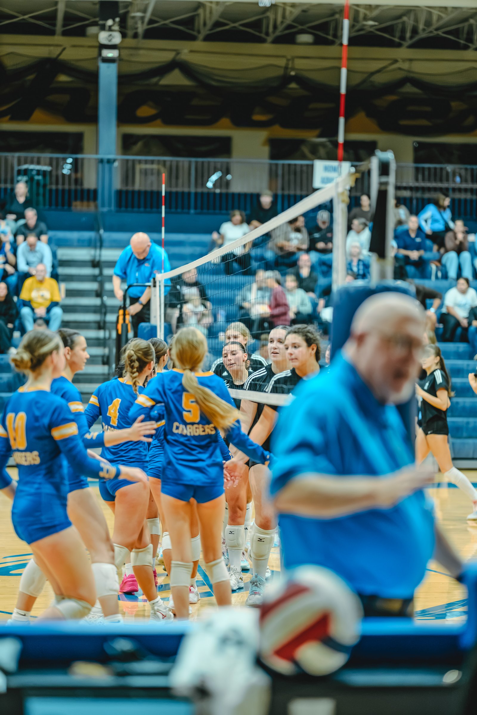 Players in blue and black jerseys huddle on the court during a volleyball game. Player 10 in blue and player 5 in blue are visible.