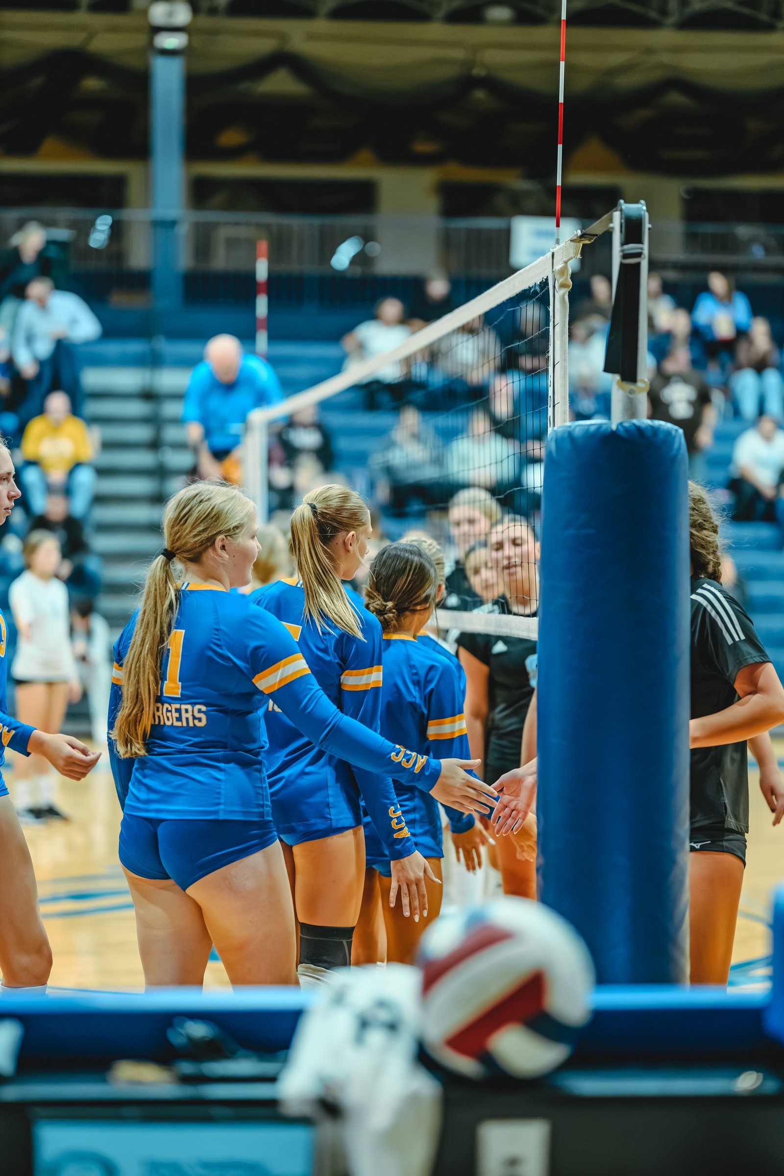 Players in blue and black jerseys shake hands at the net. Player 1 in blue has "CHARGERS" on her jersey.