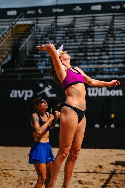 A player in a pink top and black bottoms jumps with arms extended, while a player in a blue skirt and black top watches.