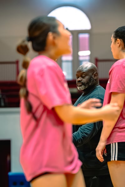 A player in a pink shirt and a player in black shorts stand near a coach in a dark sweatshirt.