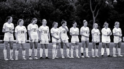 A line of players in white uniforms stand on a field, some with hands clasped, others with arms at their sides.