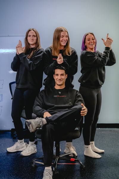 Four people in black tops pose around a chair.