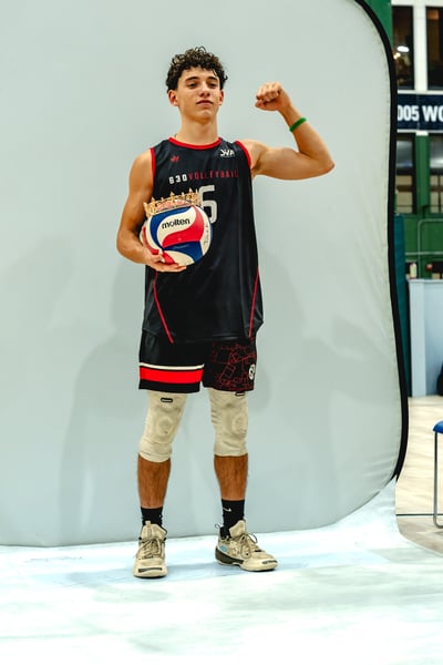 A player in a black top and shorts holds a volleyball and flexes his arm.