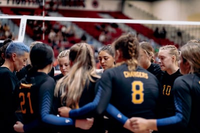 A player in a black uniform with yellow lettering stands with her arms around other players.
