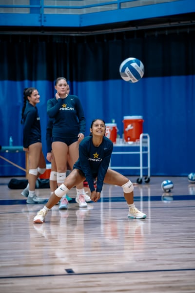 A player in a dark blue uniform crouches low with hands extended toward the floor as a volleyball flies overhead.