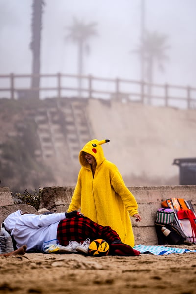 A person in a yellow Pikachu costume stands over a person lying on the sand next to a ball.