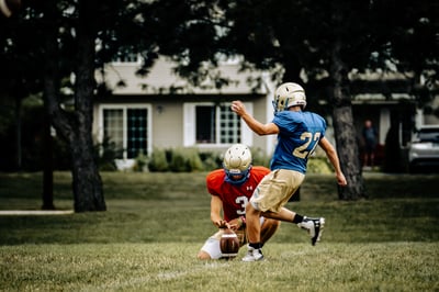 A player in red kneels with a football while a player in blue kicks the ball.