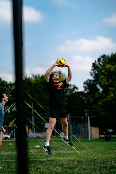 A player in black jumps with arms overhead holding a yellow ball.