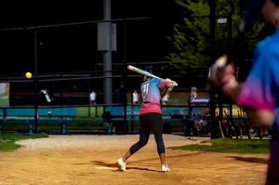 A player in a pink and blue shirt swings a bat at a yellow ball.