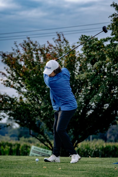 A golfer in a blue jacket and black pants swings a club at a golf ball on a tee.