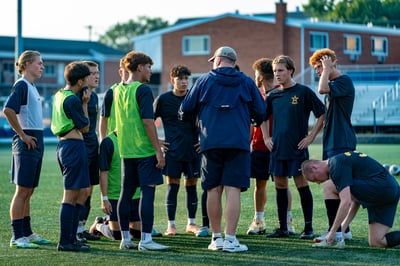 A player in a dark uniform crouches to tie his shoe while other players in dark uniforms and bright green vests stand around him.