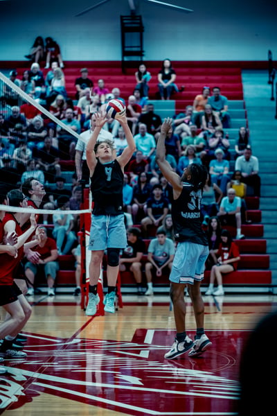 A player in black jumps and reaches overhead for the ball while a player in black reaches up to block.