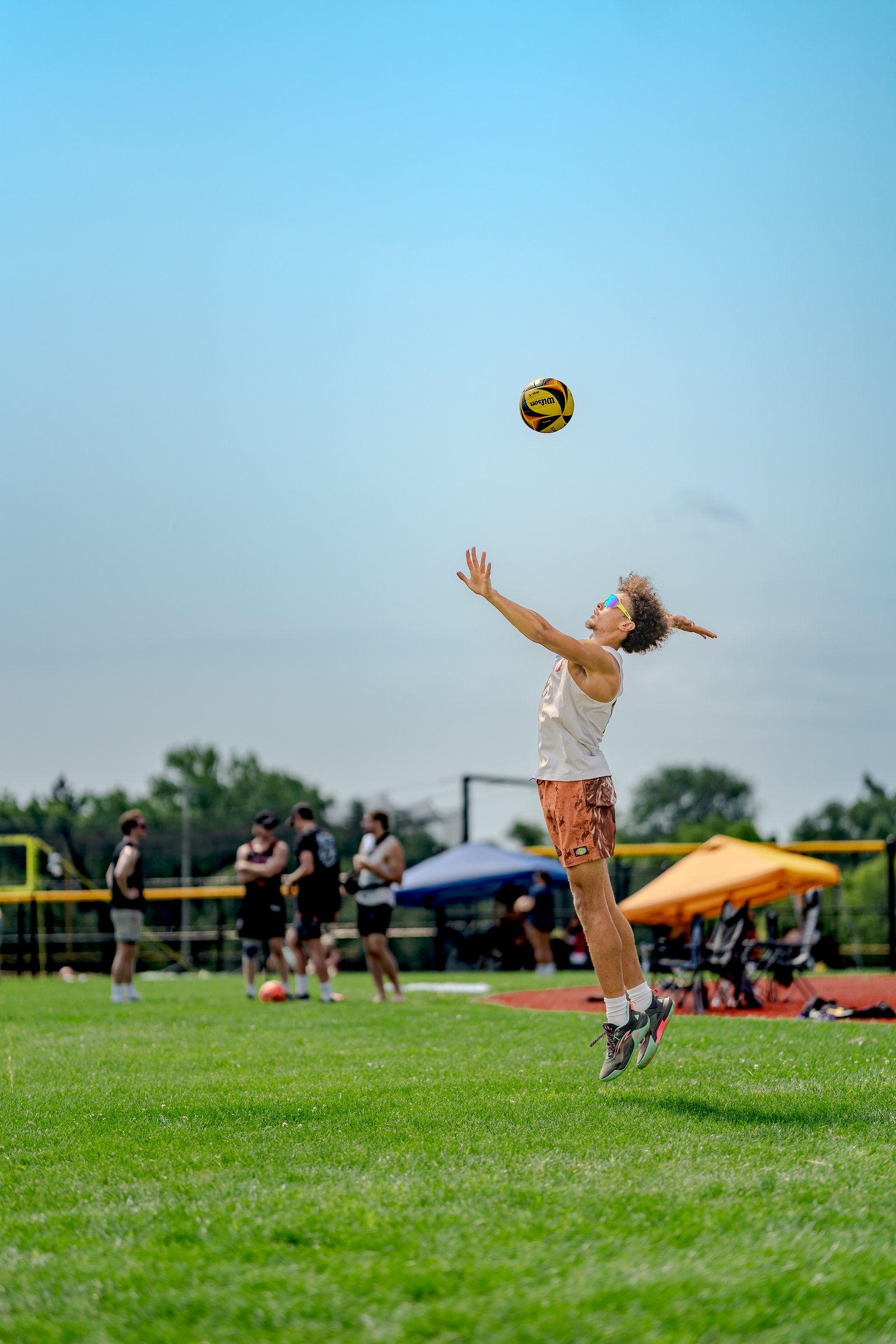 A player in a white tank top and brown shorts jumps to hit a volleyball against a clear blue sky.