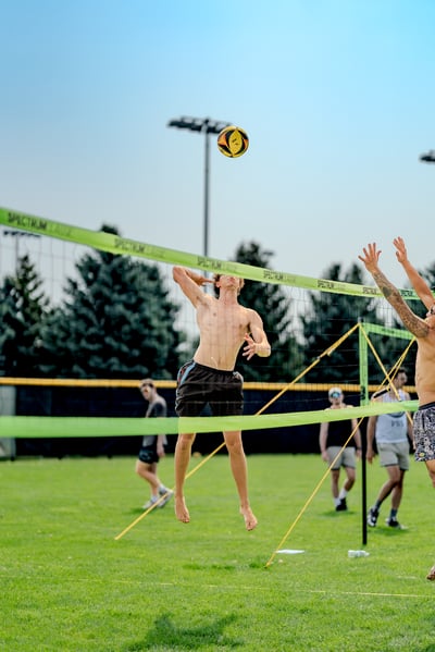 A player in black shorts jumps to hit a volleyball over the net while another player in patterned shorts reaches up.