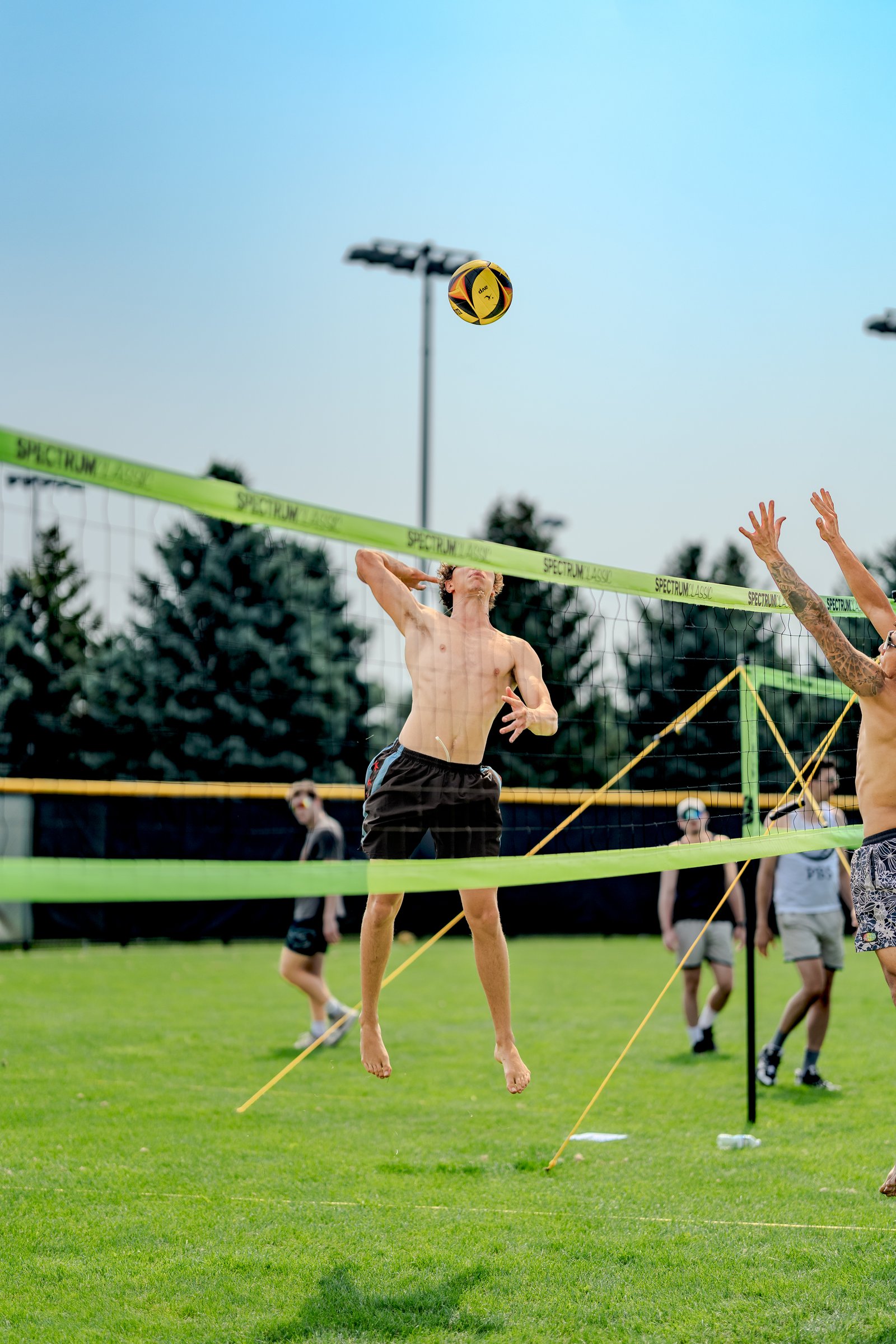 A player in black shorts jumps to hit a volleyball over the net while another player in patterned shorts reaches up.