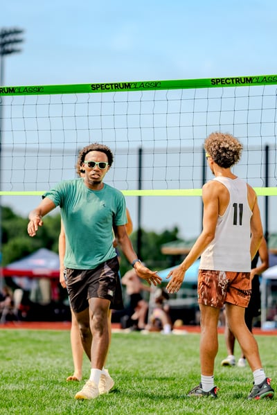Two players in green and white tops high-five near a volleyball net on a sunny day.
