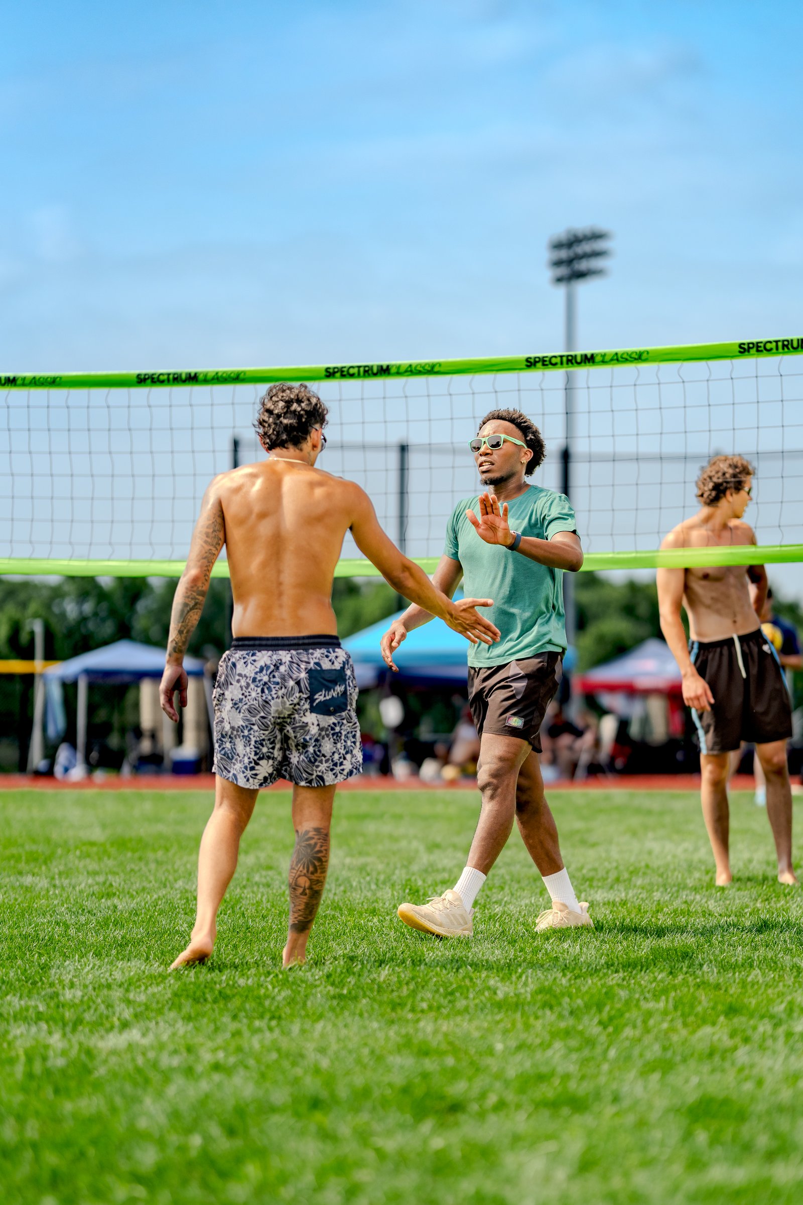 Two players in green and patterned shorts interact near a volleyball net on a grassy field under a blue sky.