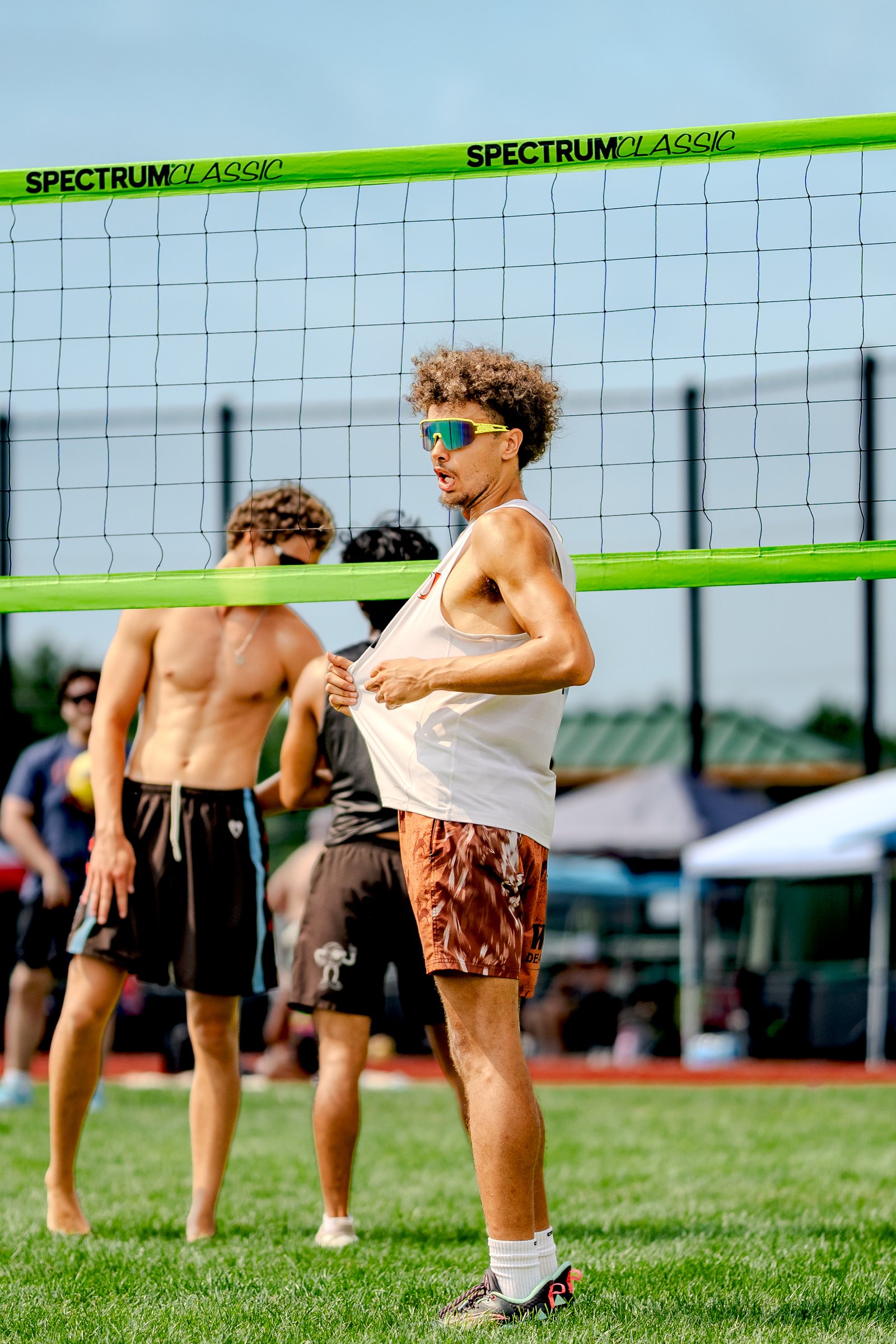 A player in a white tank top and brown patterned shorts adjusts his shirt near the net, with other players in black shorts and a blue-striped shirt visible.