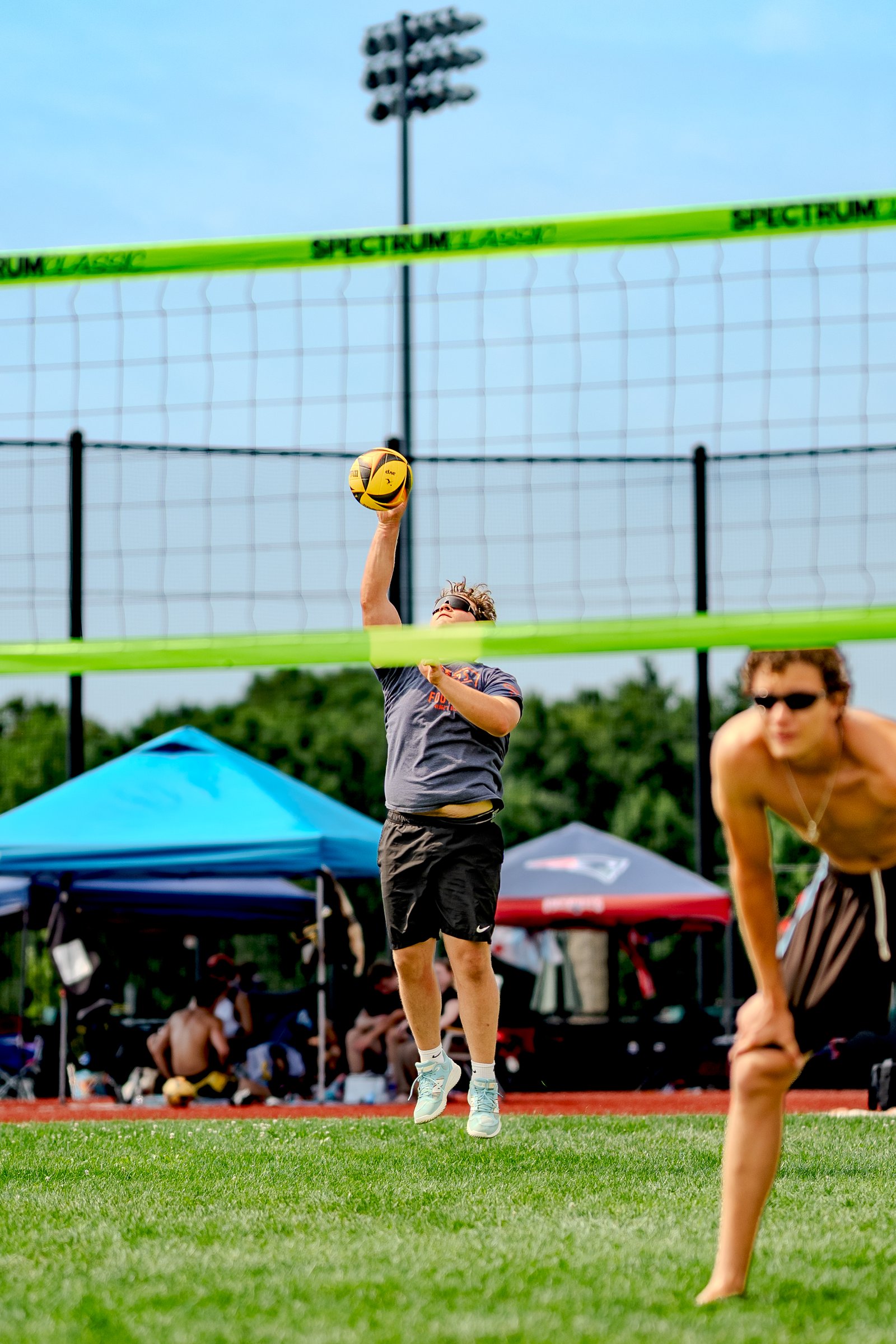 A player in a gray shirt and black shorts jumps to serve a volleyball over the net, with another player in black shorts in the foreground.