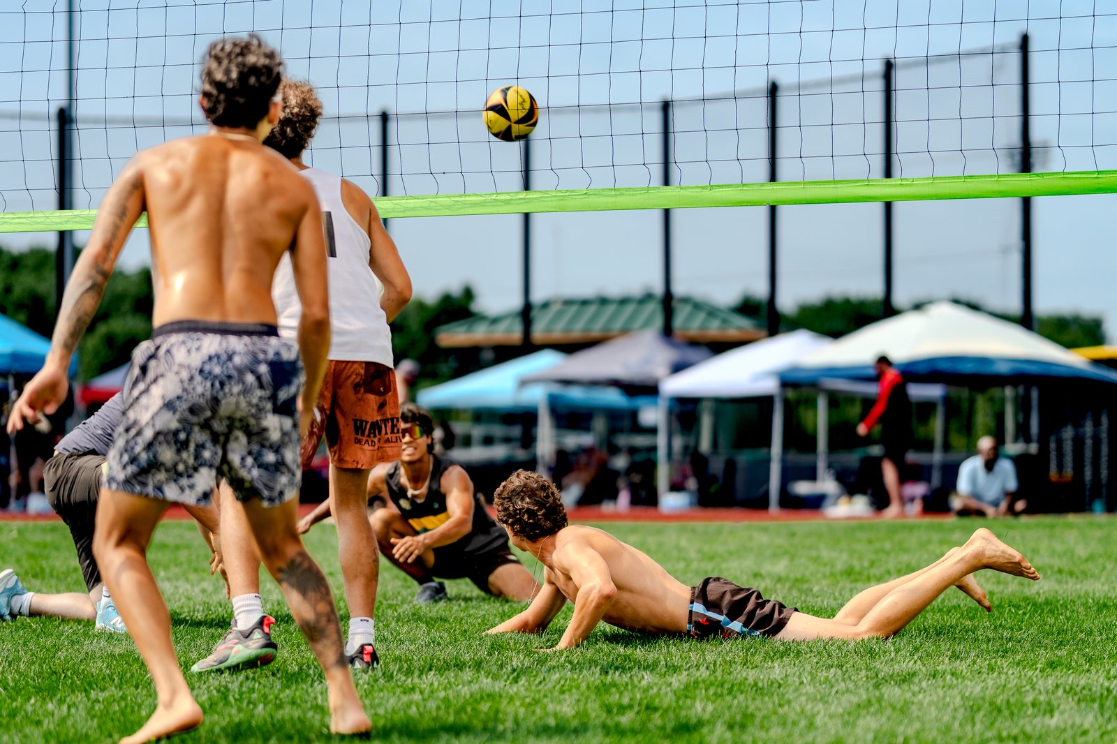 Players in black and white tops and patterned shorts dive for a volleyball on a grassy court.