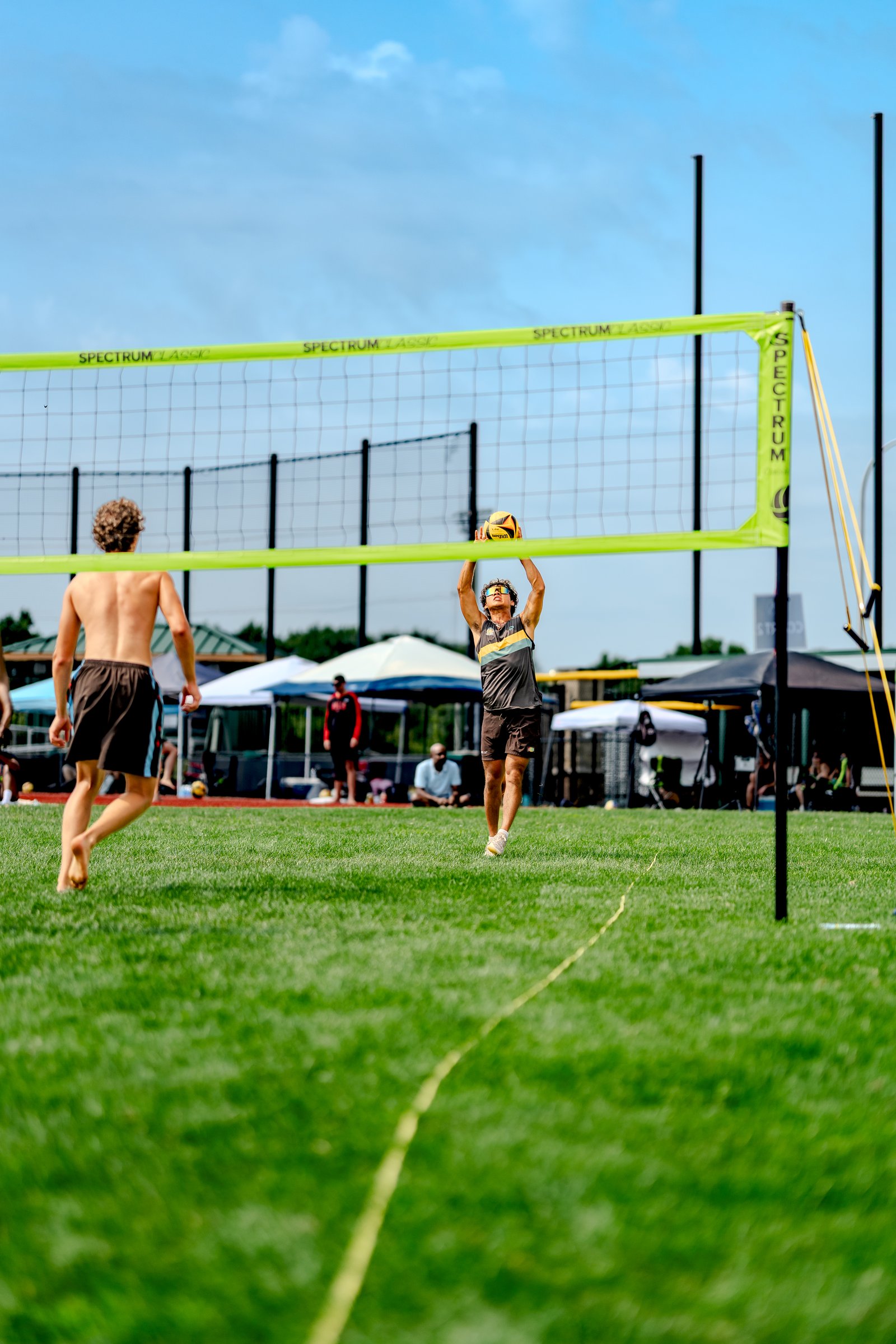 A player in a gray and yellow top sets a volleyball on a grassy court with a player in brown shorts in the foreground.