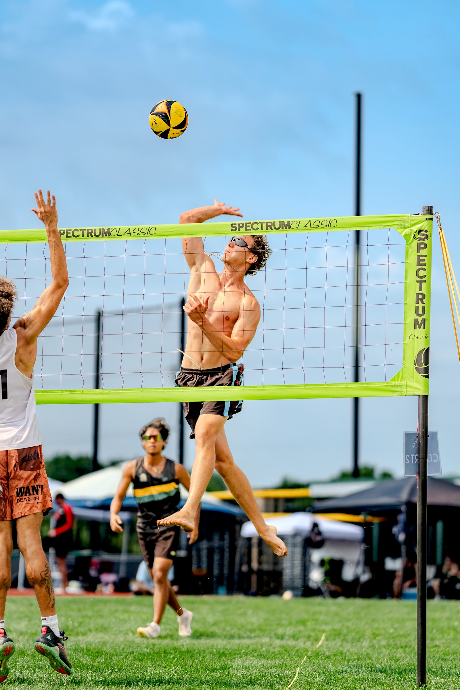 A player in black shorts jumps to spike a volleyball over the net, with a player in a white top reaching to block.