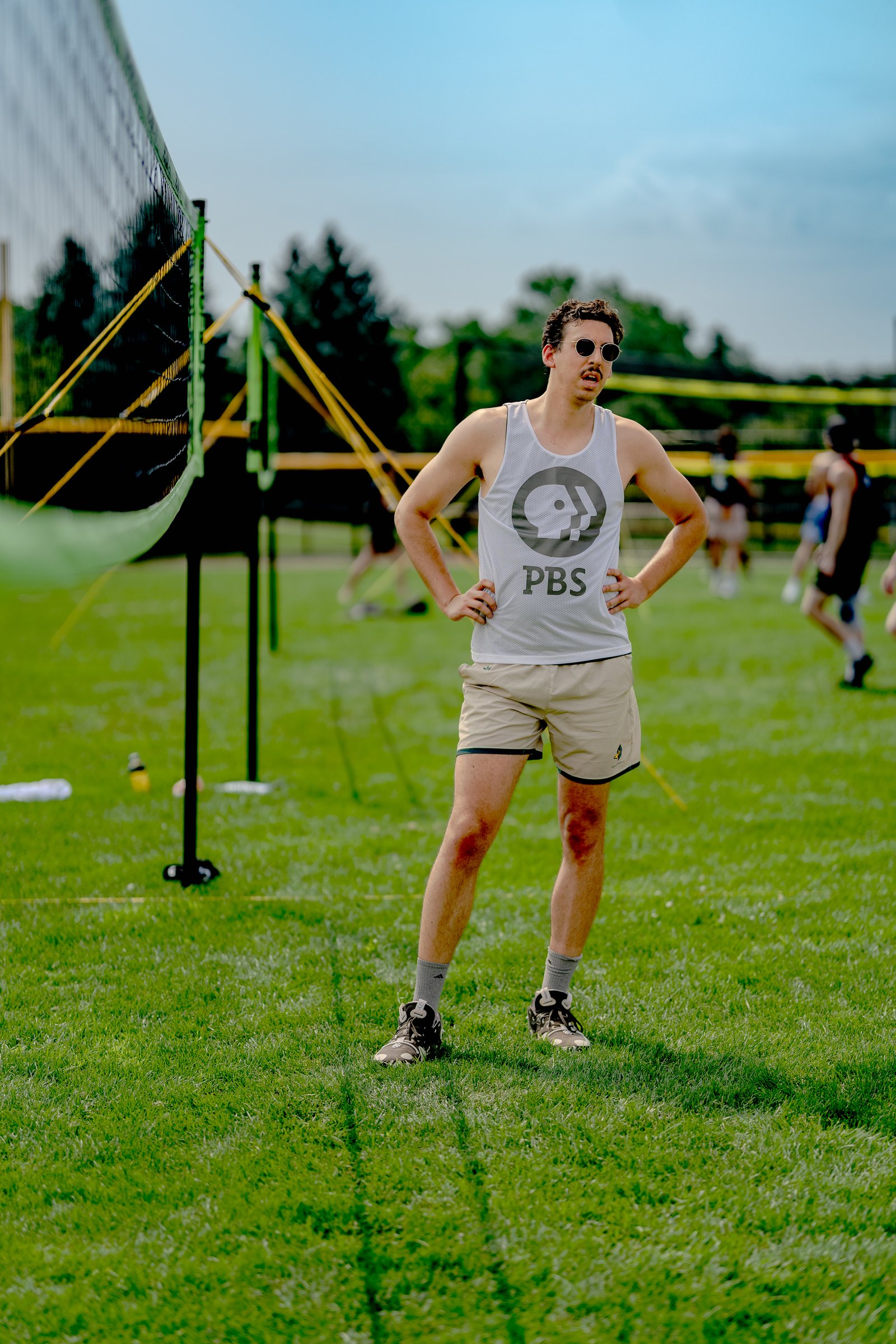 A player in a white tank top and tan shorts stands on the grass court with hands on hips, facing away from the net.
