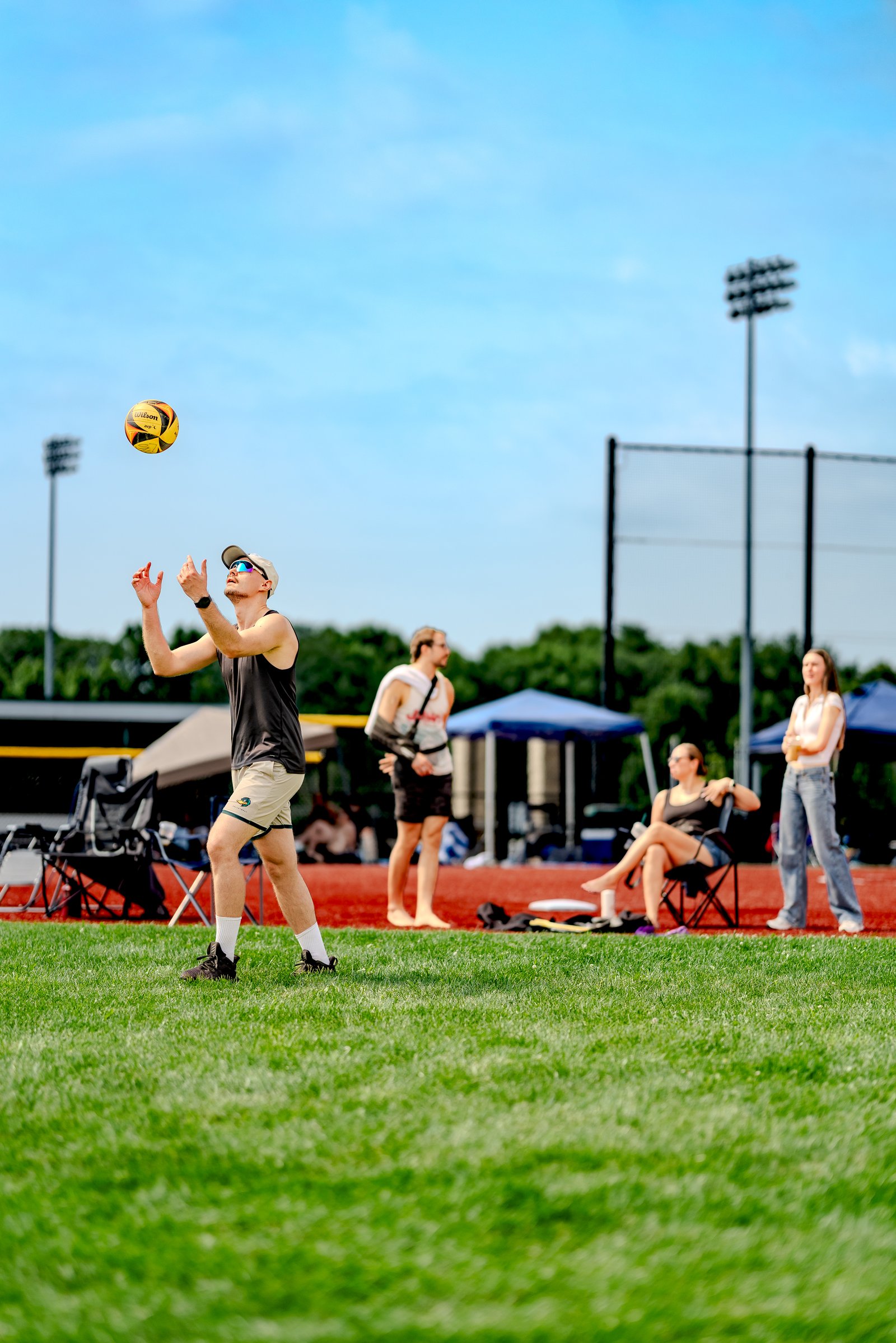 A player in a black tank top and tan shorts tosses a volleyball in the air on a grassy court with other players watching.