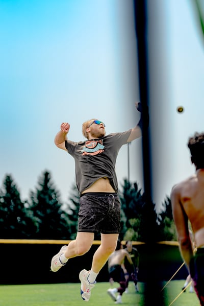 A player in a dark t-shirt and black shorts jumps to hit a volleyball, with other players visible in the background.
