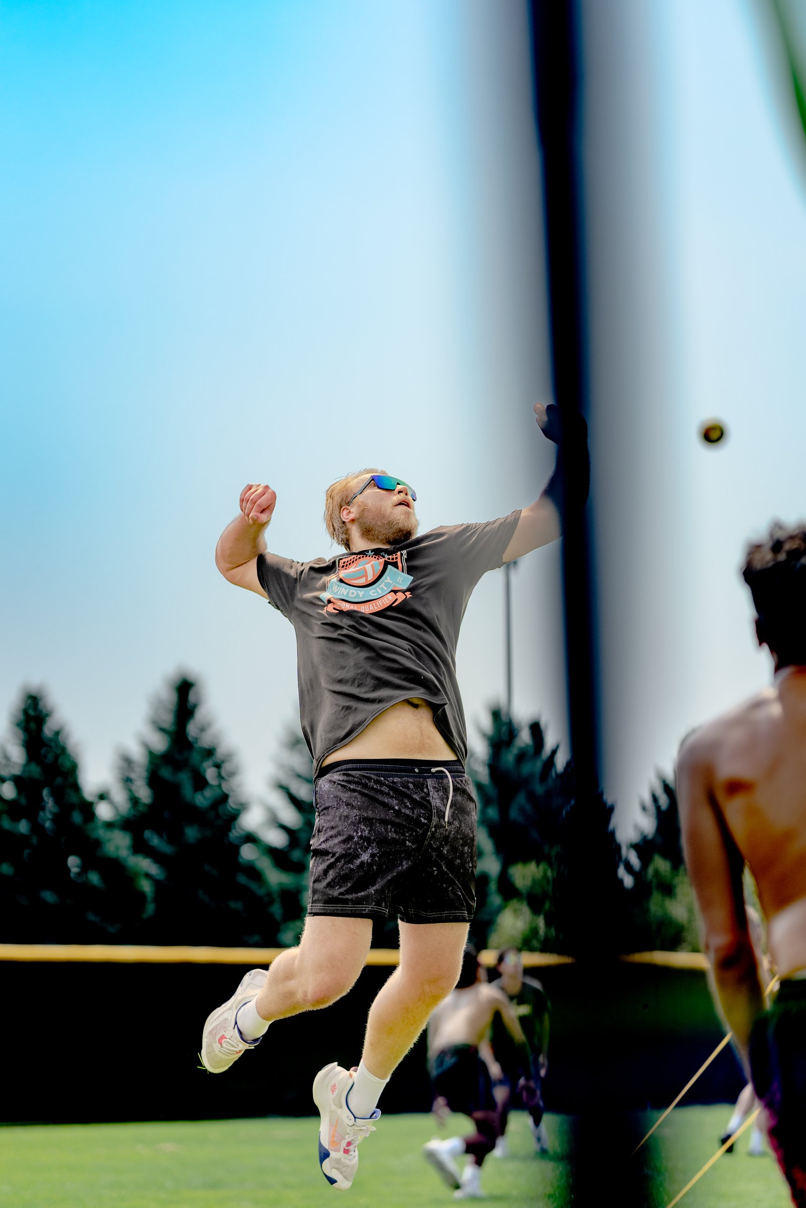 A player in a dark t-shirt and black shorts jumps to hit a volleyball, with other players visible in the background.