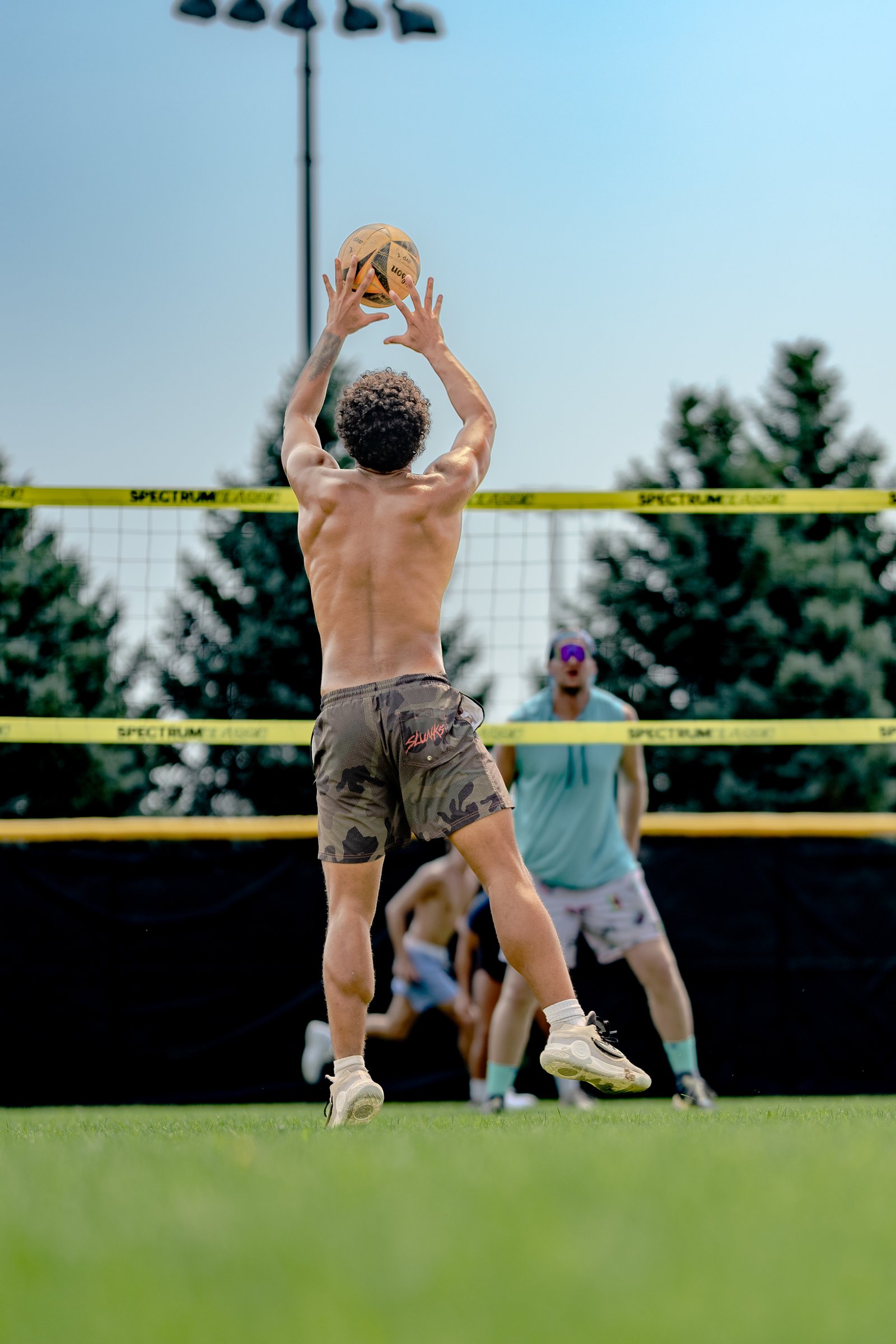 A player in camouflage shorts prepares to set a volleyball over the net, with other players in blue and white visible on the court.