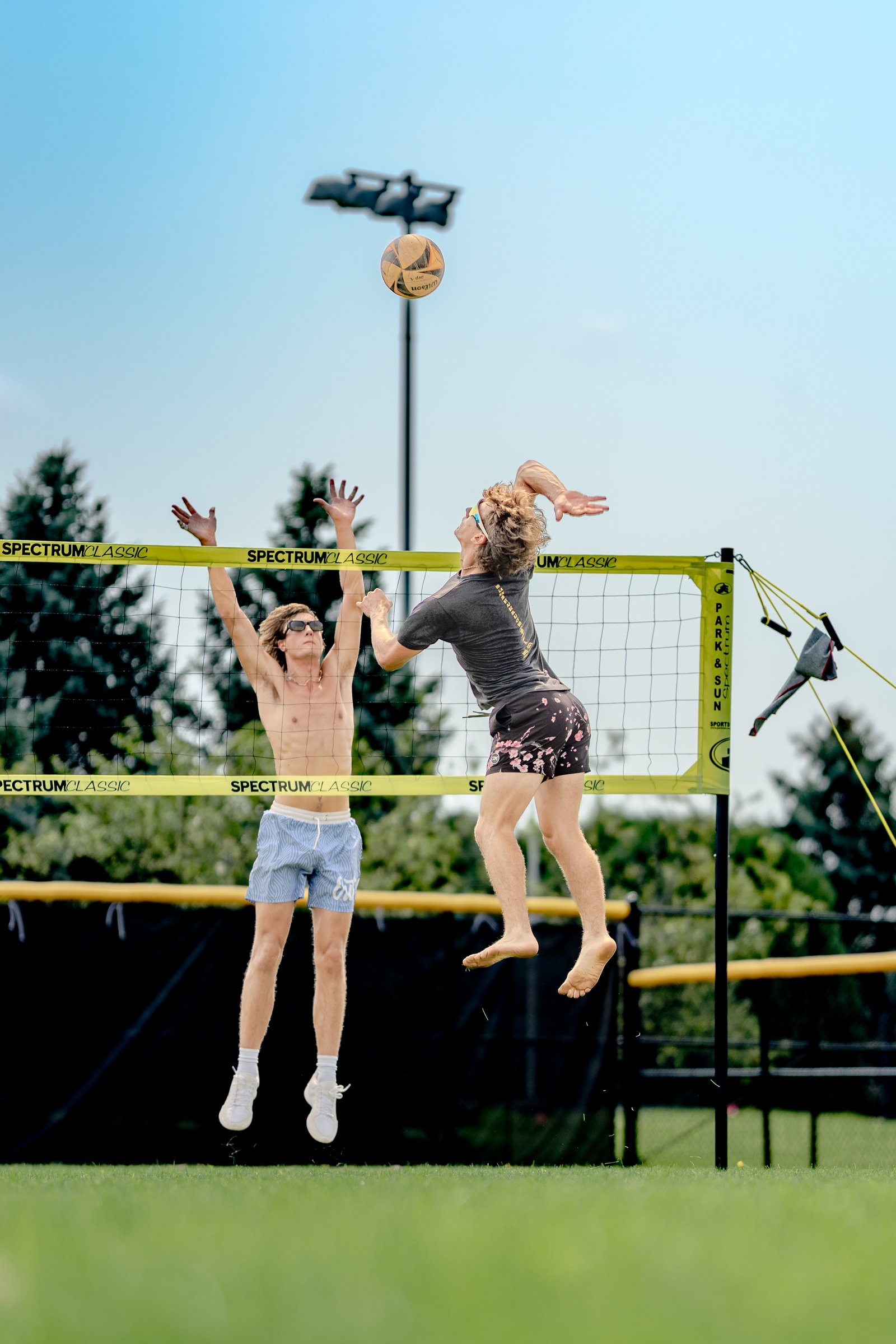 Two players jump at the net, one reaching for the ball, with a volleyball net and trees in the background.