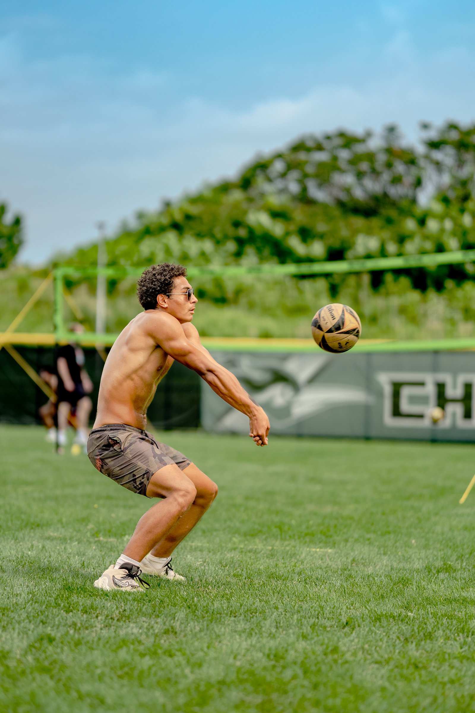 A player in camouflage shorts digs a volleyball on a grassy field with a blurred player in black in the background.