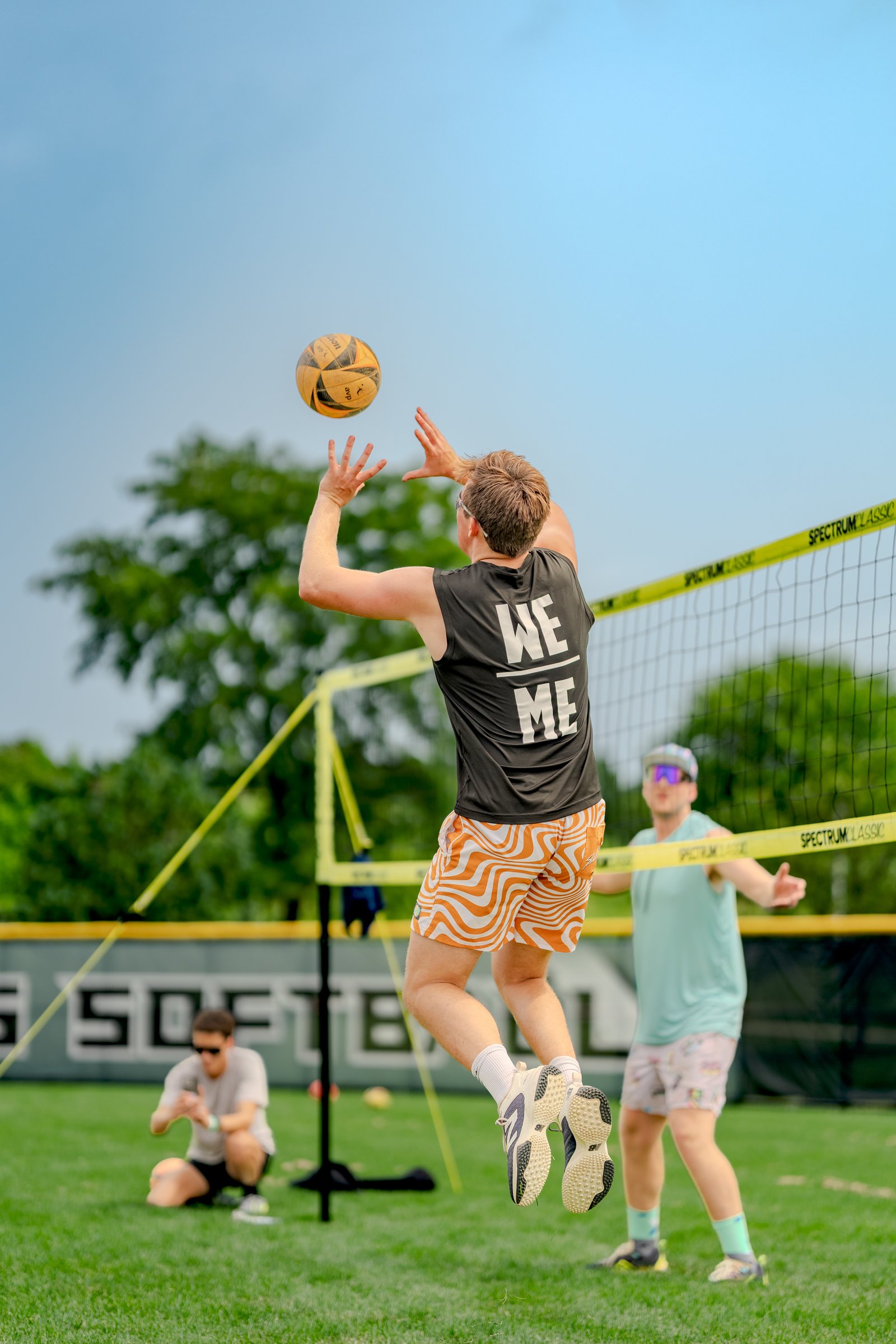 A player in a black top with "WE ME" on the back jumps to hit a volleyball, with players in teal and gray visible on the court.