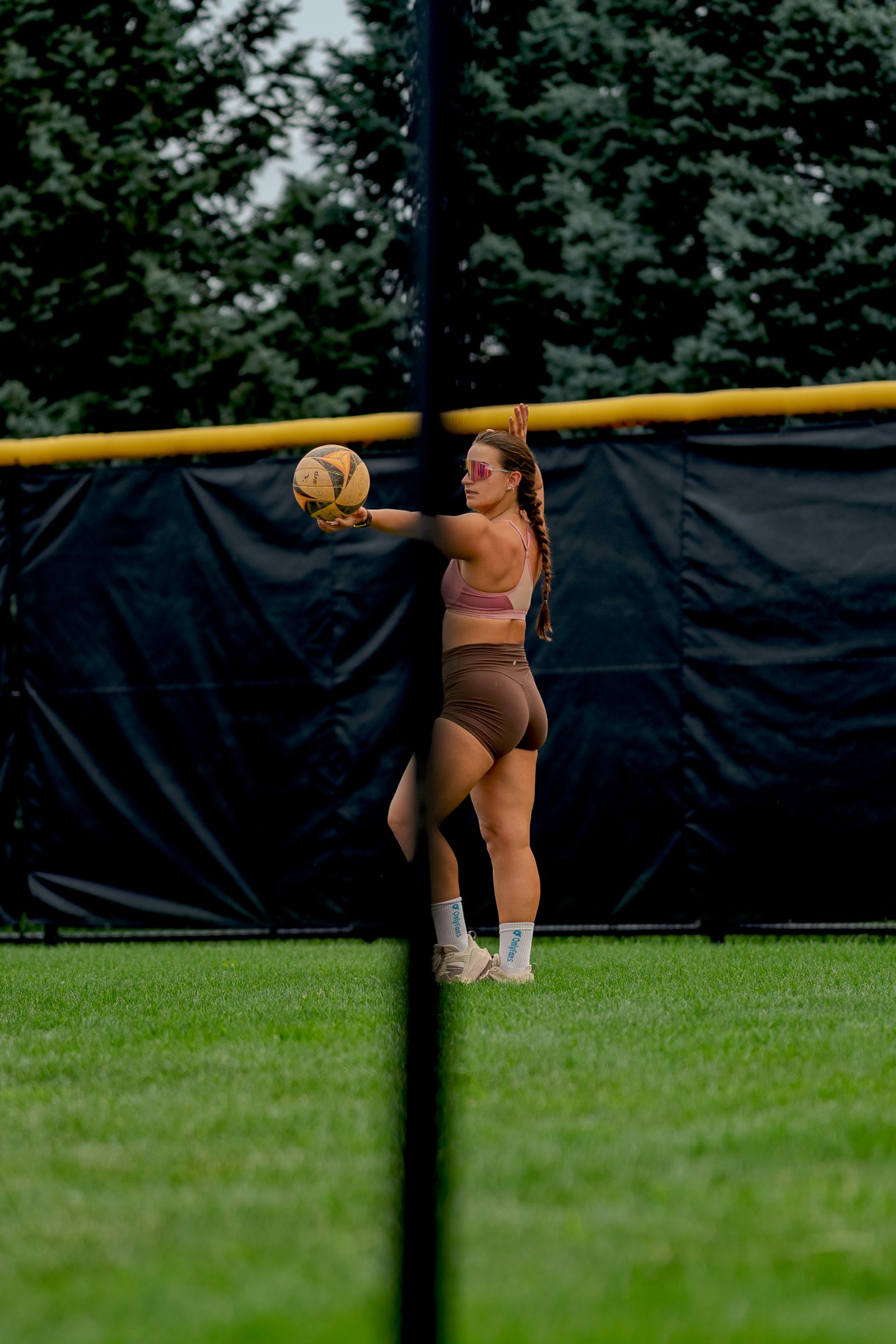 A player in brown prepares to serve a volleyball on a grassy court with a black net and trees in the background.