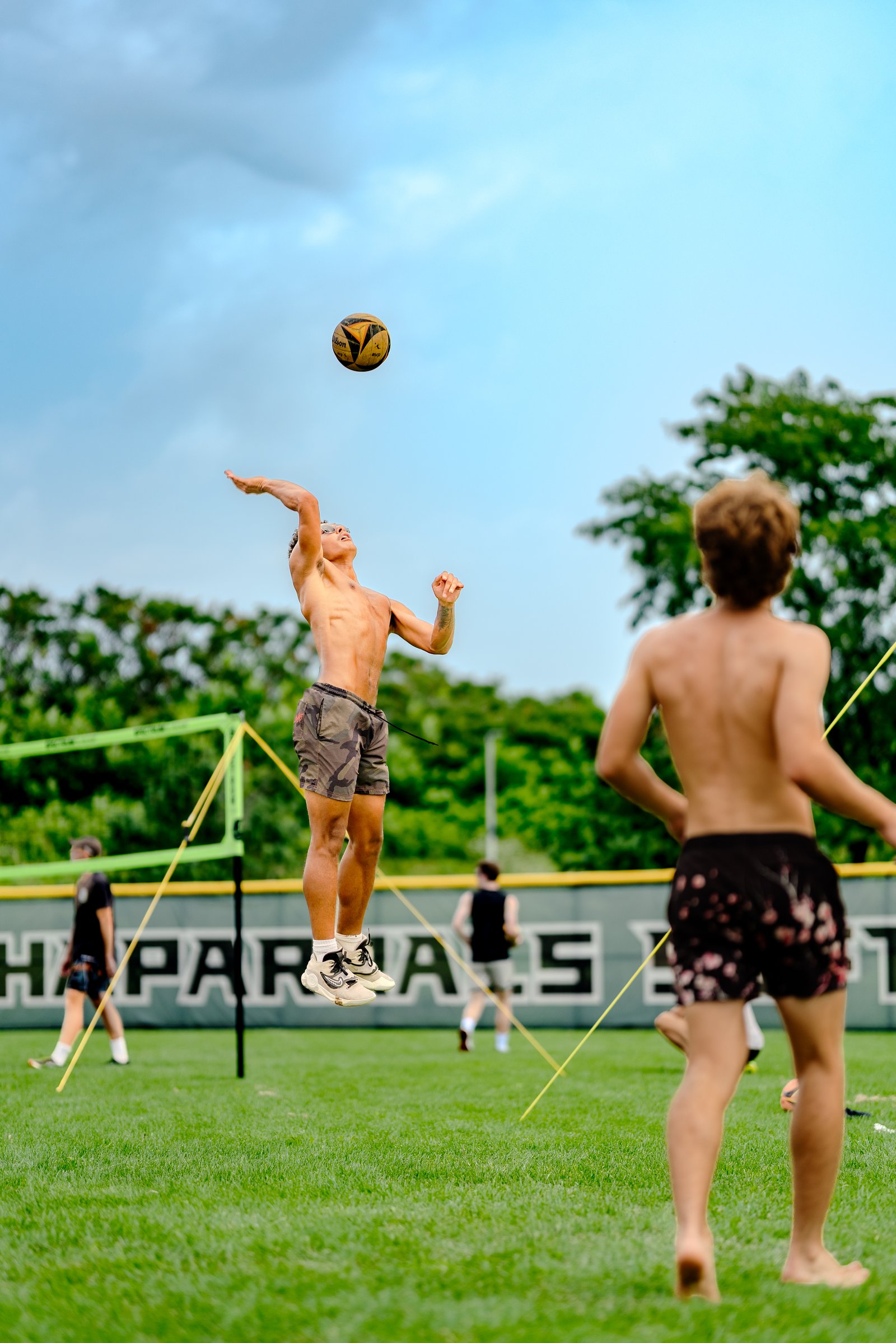 A player in camouflage shorts jumps to hit a volleyball, with other players on the grass court and a green net in the background.