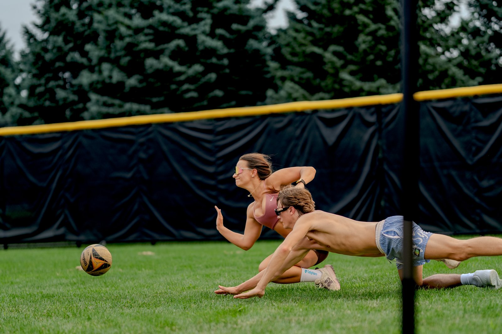 Two players dive for a volleyball on a grassy court with a black fence and trees in the background.