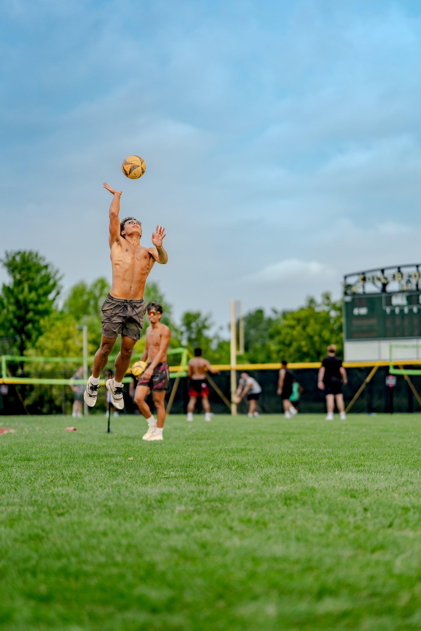 A player in gray shorts jumps to hit a volleyball over the net, with other players in the background on a grassy field.