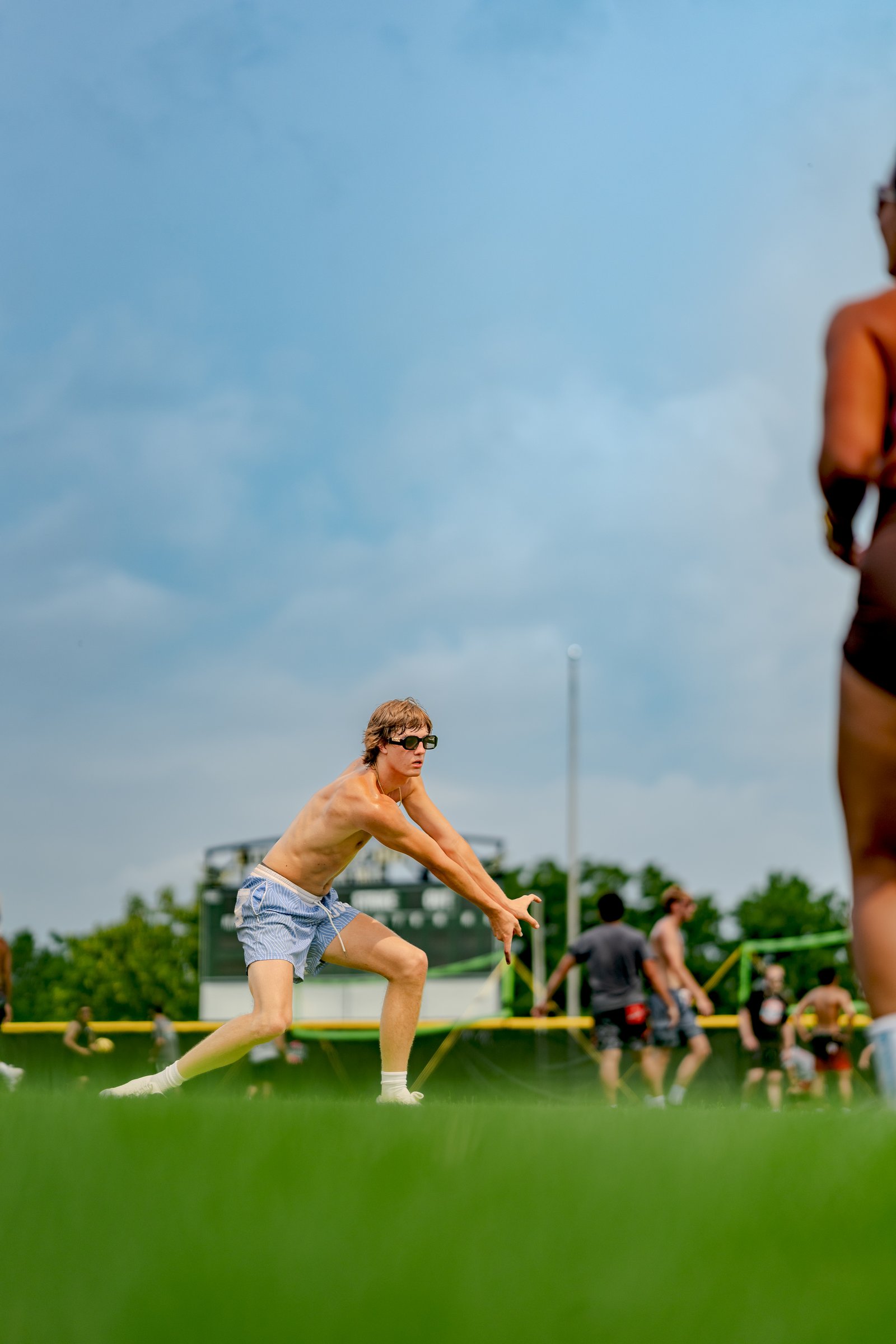 A player in blue shorts prepares to dig a volleyball on a sunny day with a scoreboard in the background.