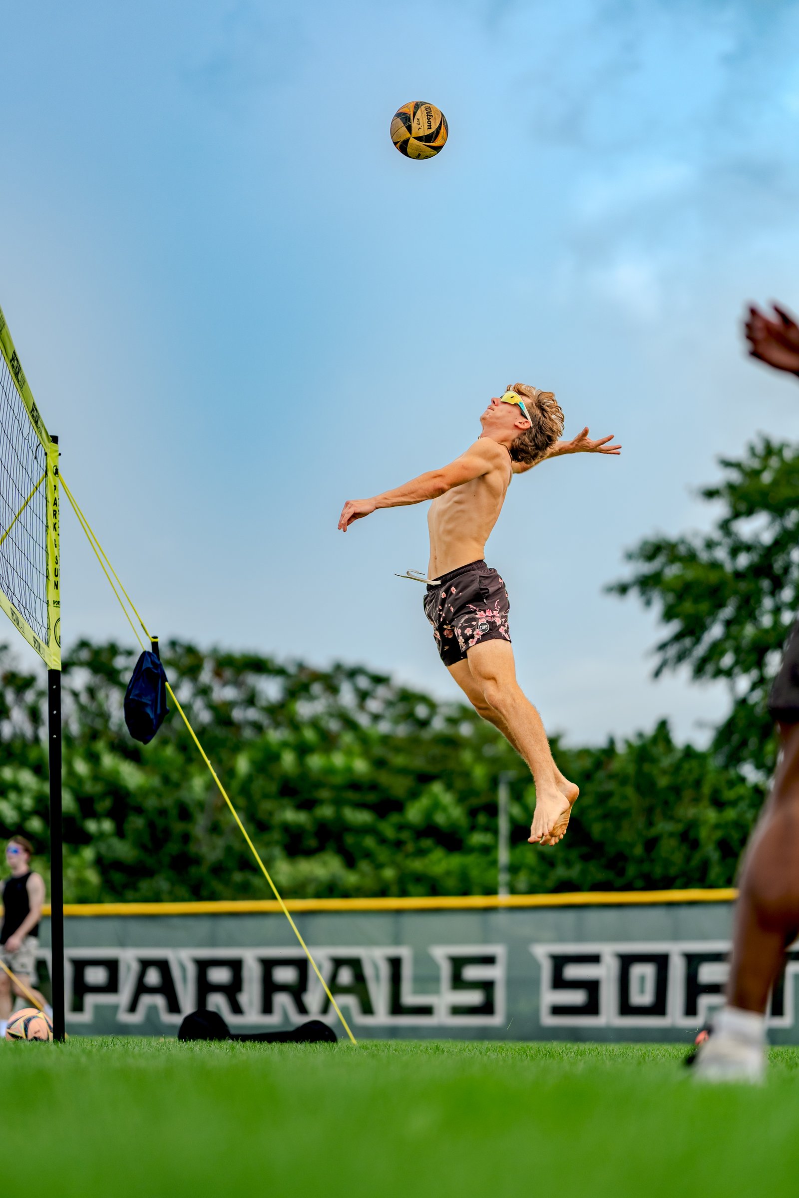 A player in black floral shorts jumps to hit a volleyball against a blue sky with trees in the background.