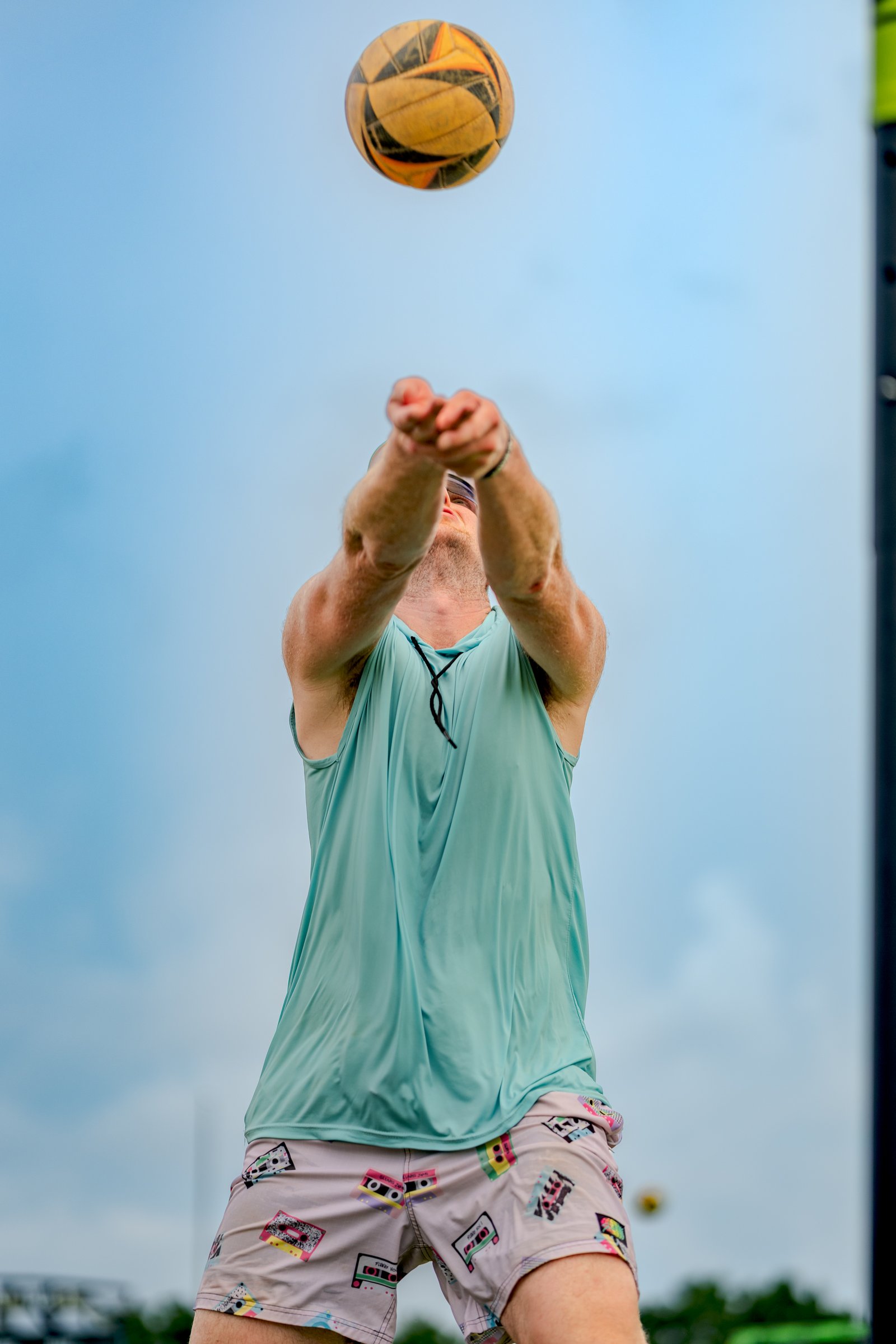 A player in a teal top and patterned shorts sets a volleyball against a cloudy sky.