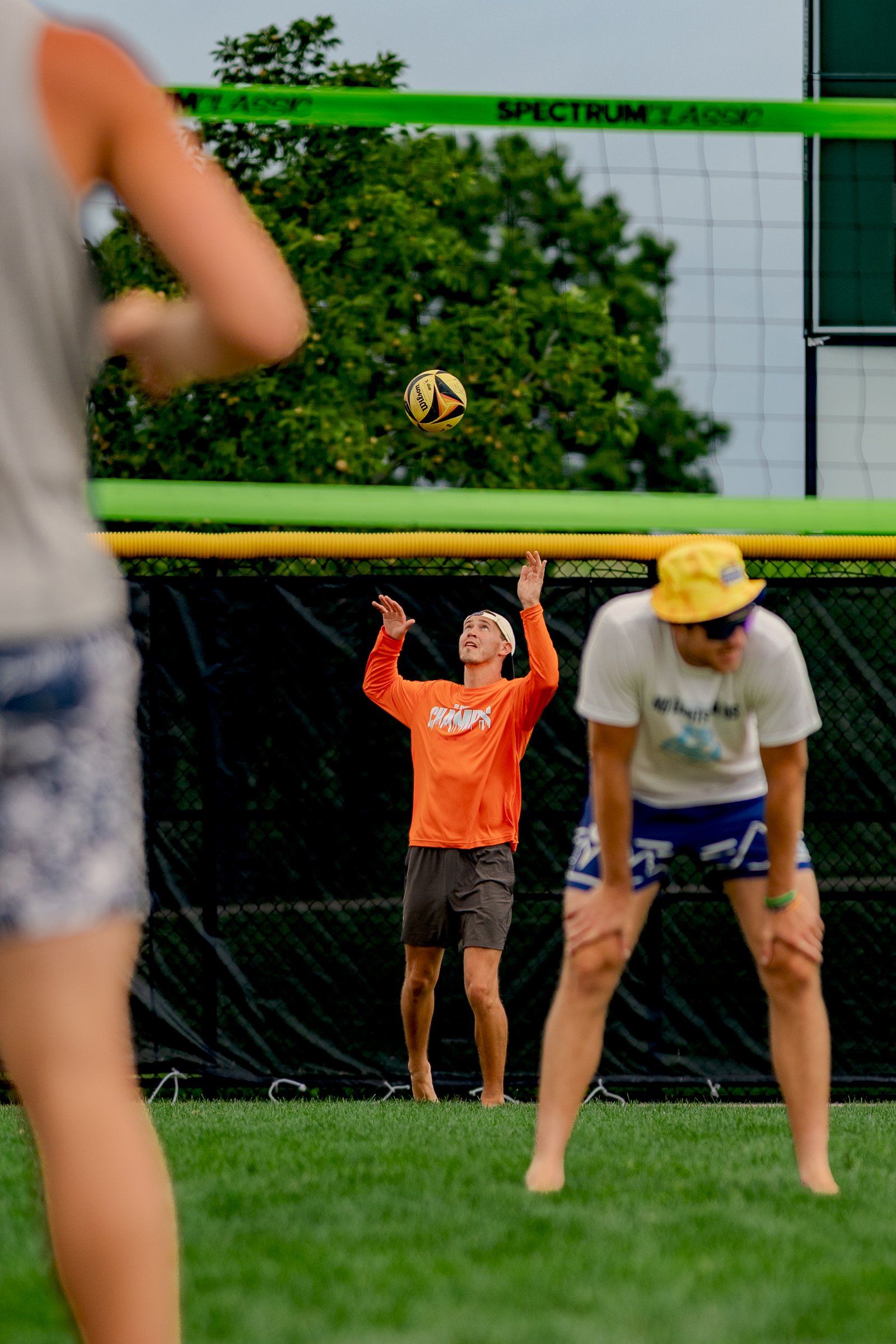 A player in orange prepares to set a volleyball as two other players in white and blue shorts watch.