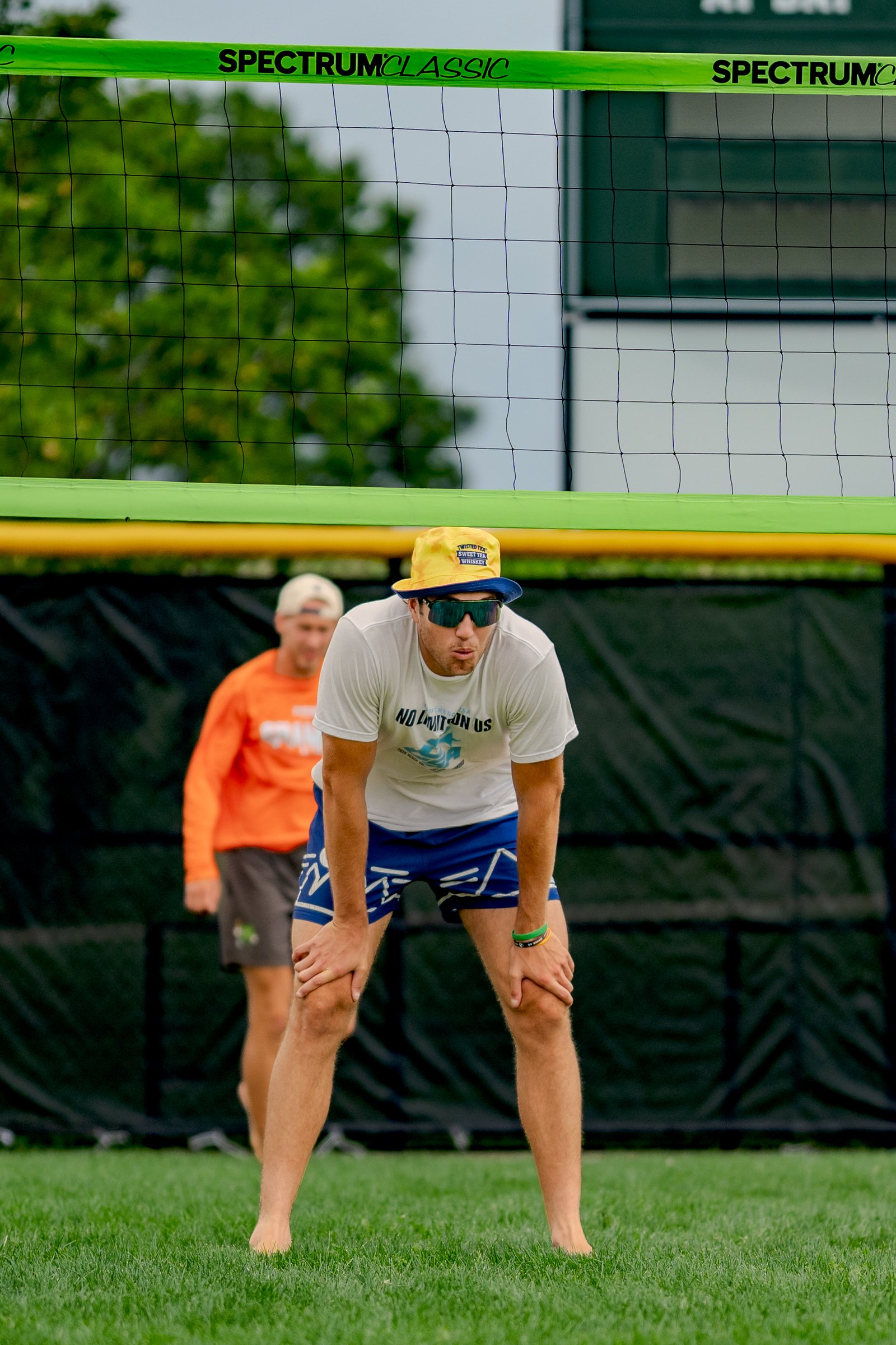 A player in blue shorts and a white shirt bends over on the grass, while another player in an orange shirt stands behind him.