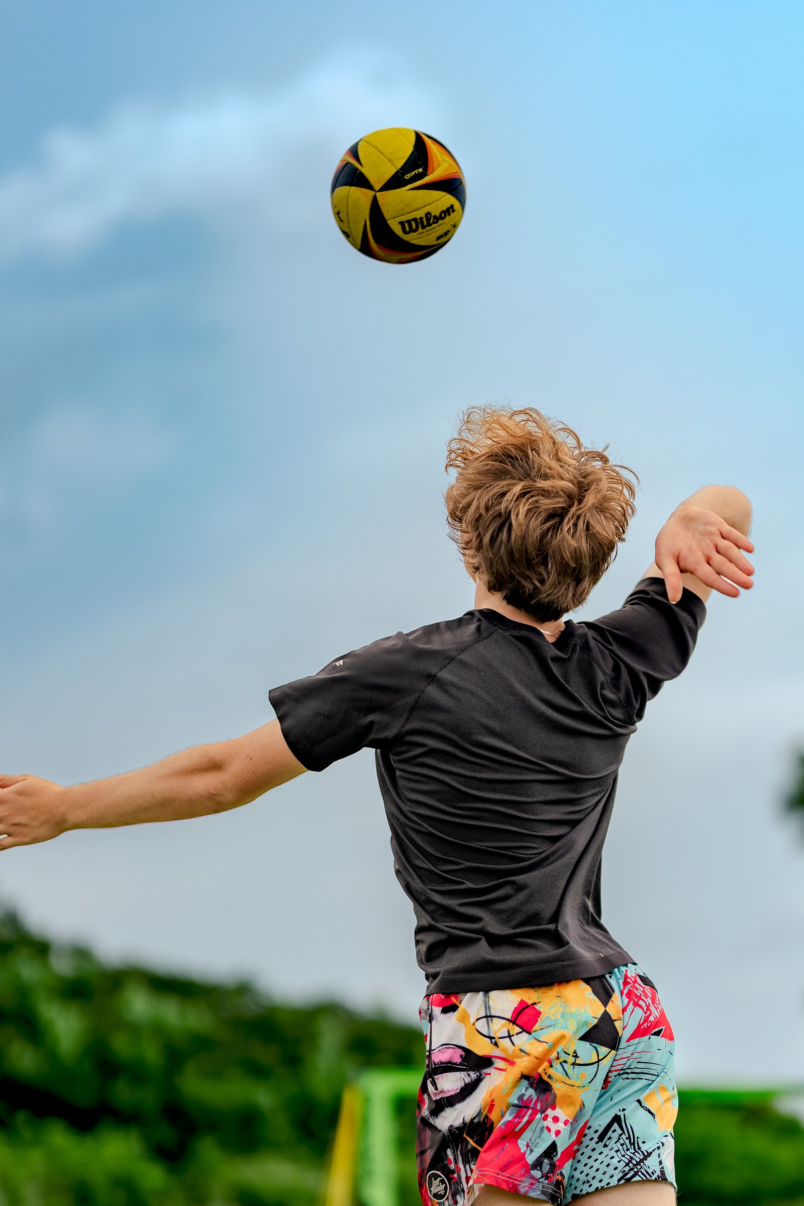 A player in a black top and colorful shorts serves a yellow and black volleyball against a cloudy blue sky.