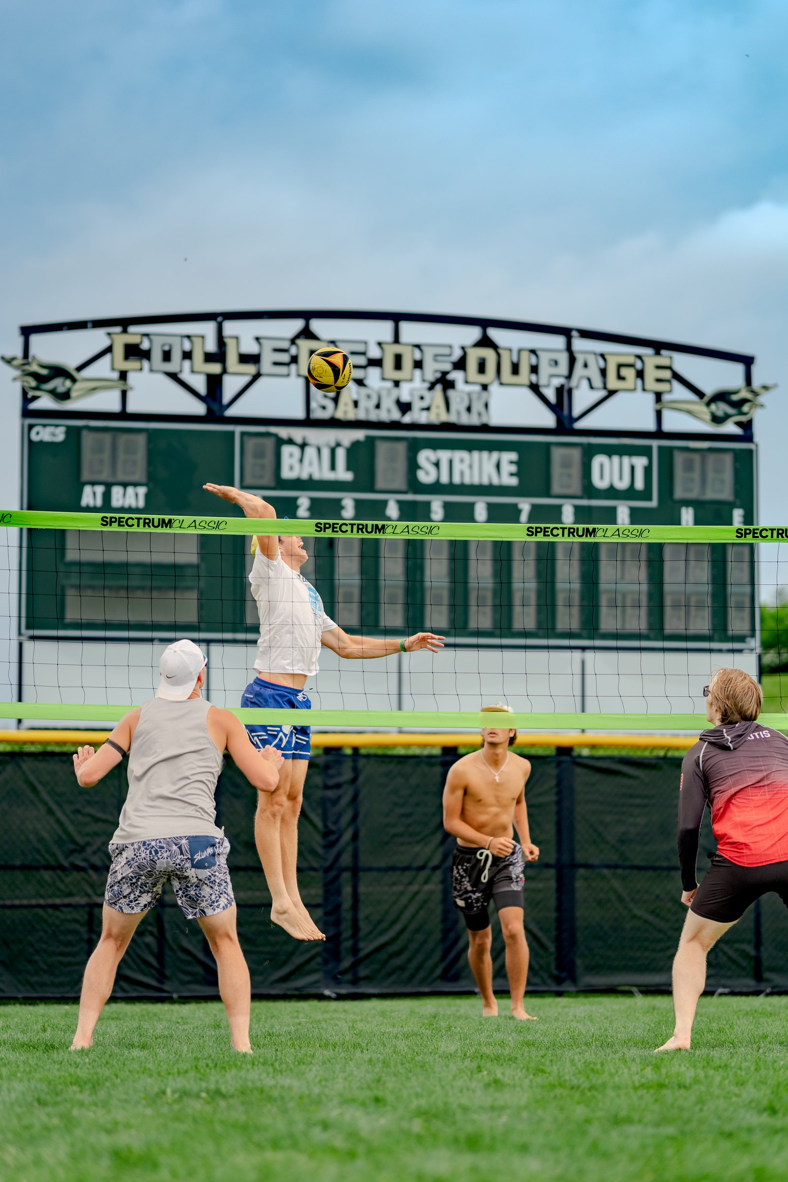 A player in a white shirt jumps to hit the ball over the net, with players in gray and red tops watching, under a scoreboard.