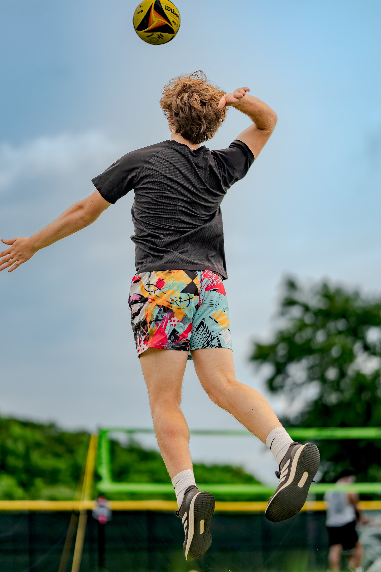 A player in a black top and colorful shorts jumps to hit a volleyball against a blue sky with trees and a net in the background.