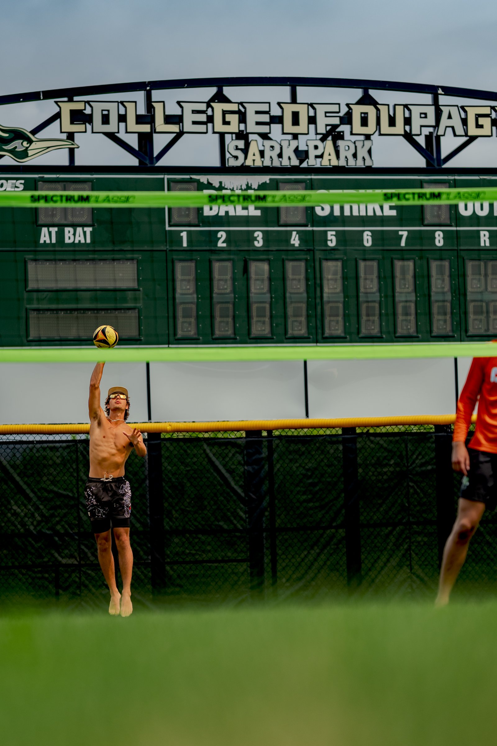 A player in black shorts jumps to hit a volleyball in front of a scoreboard at College of DuPage Sark Park.