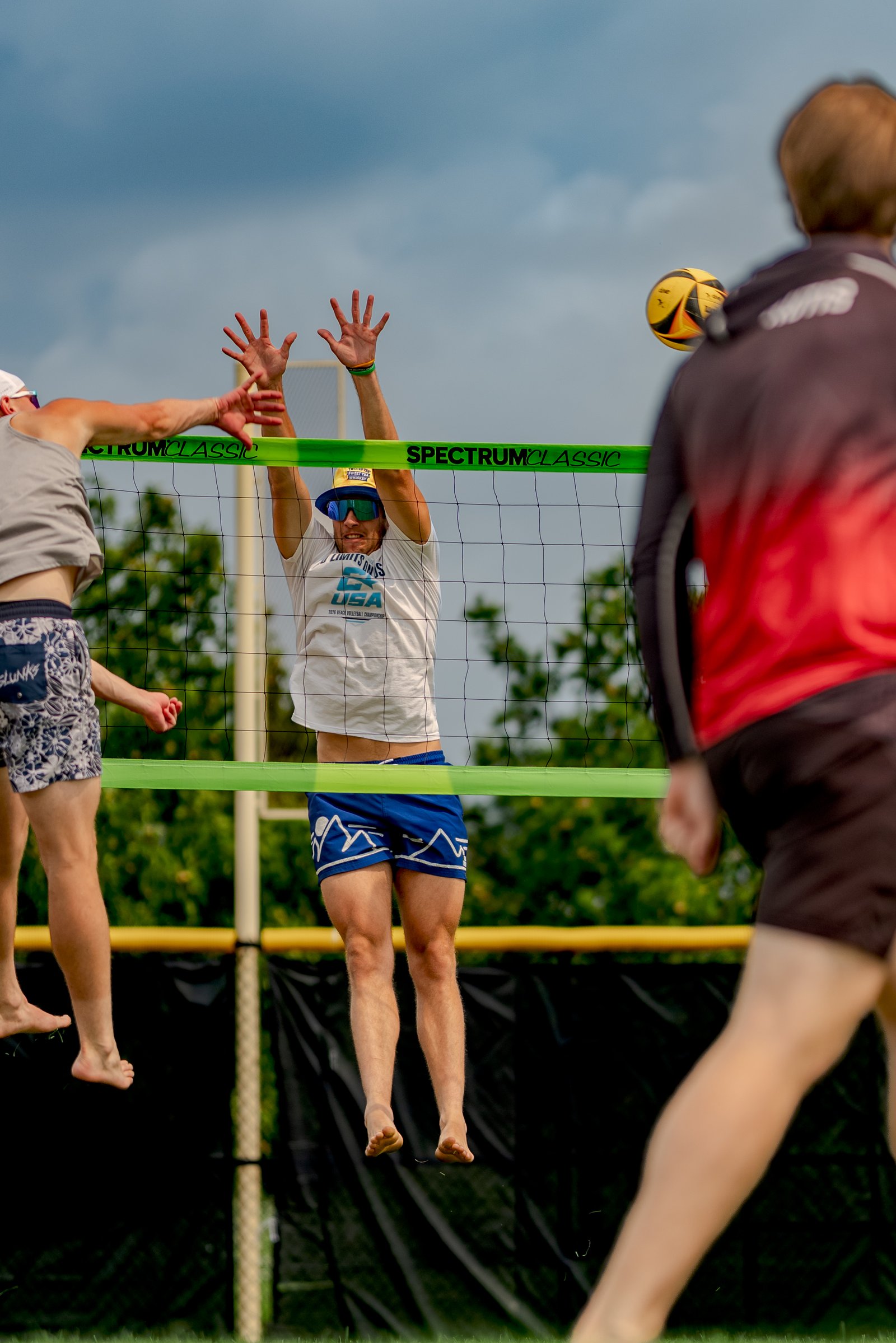 Players in white and blue shorts jump at the net during a volleyball game, with a ball in the air.