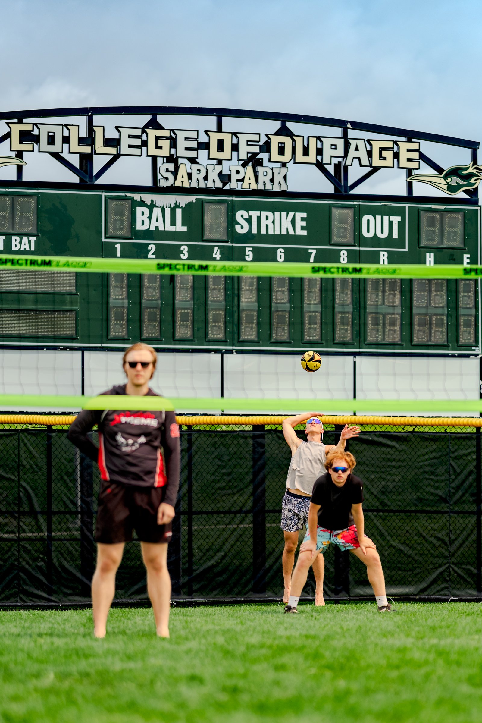 Players in black, gray, and patterned shorts play volleyball near a scoreboard at Sark Park.